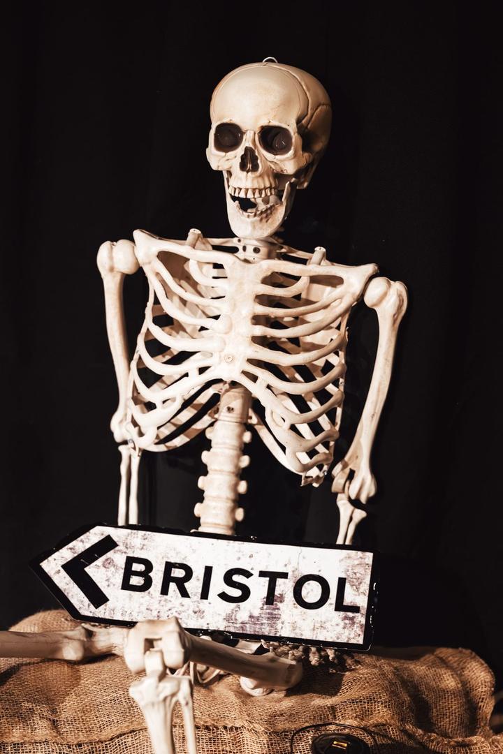 A plastic skeleton sits among burlap sacks holding a wooden sign pointing to Bristol