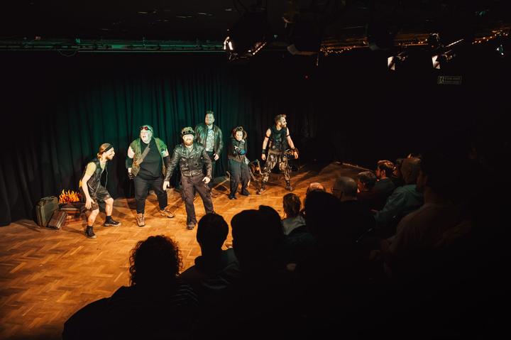 The cast in wasteland costume spread across a dark stage, seen from the audience