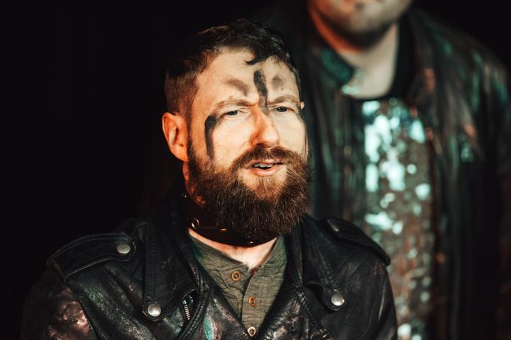 Close-up of a bearded performer with black face paint, a leather jacket and a buckled choker