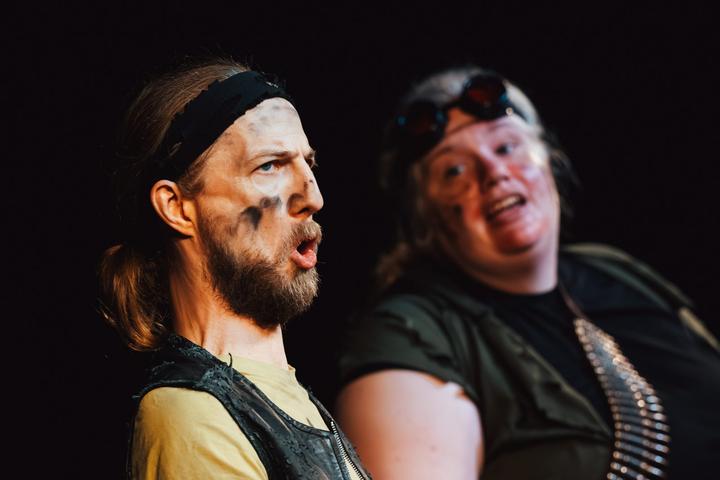 Two performers mid-scene: a man with a headband speaking and a woman with red goggles looking wide-eyed