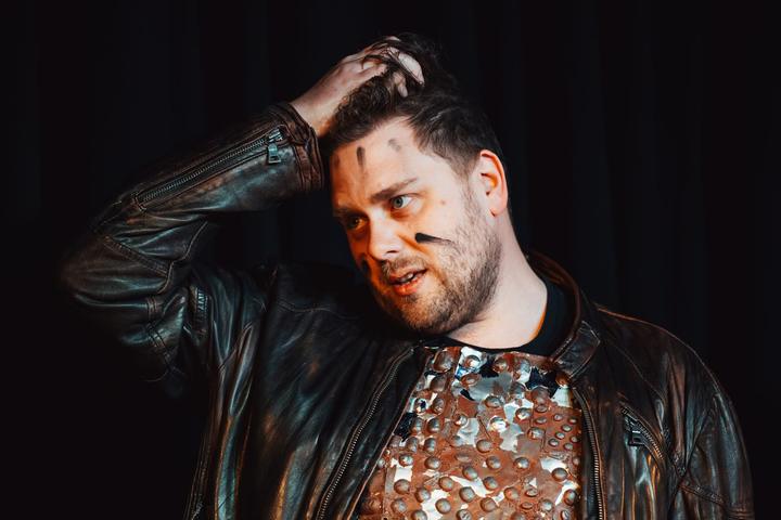 A performer in a studded leather jacket looks down, a hand in his hair