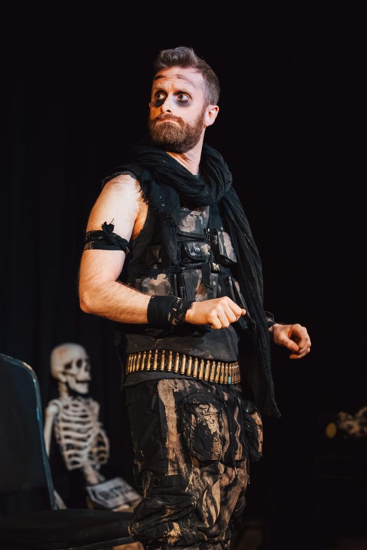 A bearded performer in a bullet belt and ripped trousers stands on stage beside a skeleton