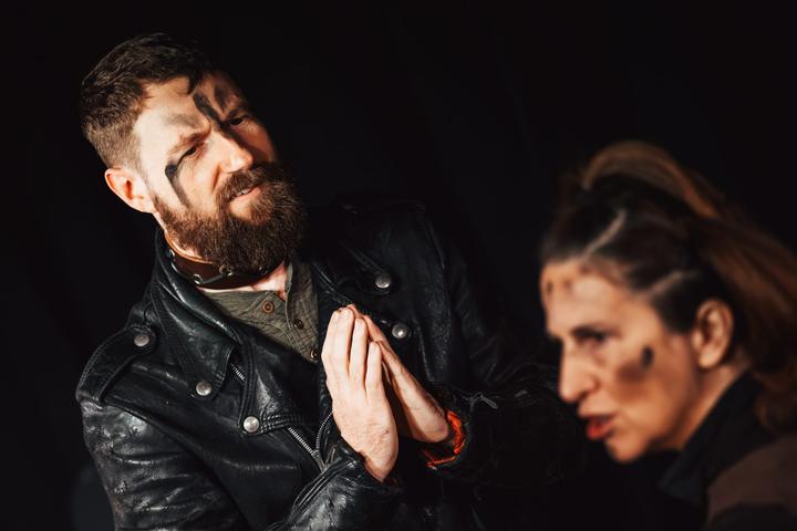 A bearded performer clasps his hands as if in prayer while another looks on