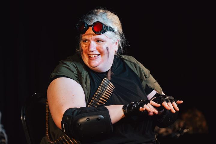 A performer with grey hair, red goggles and an ammunition belt smiles at the camera